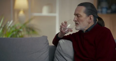 Older man sitting on sofa holding a glass of red drink in a quiet living room Видео 336287330