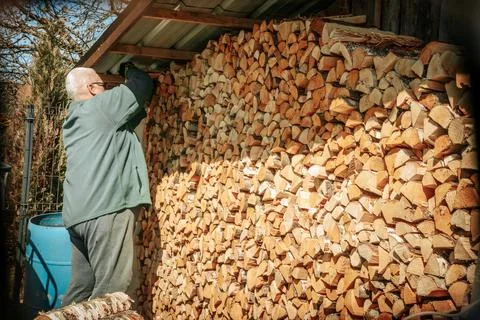 An older man stacks split firewood in a large woodpile. Photos