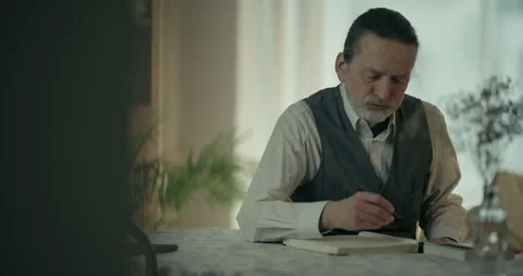 Older man writing notes at a table in a softly lit home interior Vídeos de archivo 336289459