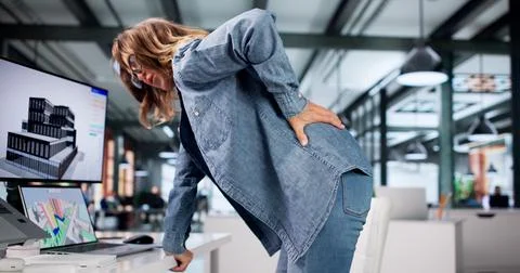 Older Woman Adjusts Posture While Working At Her Office Computer Desk. Stock Photos