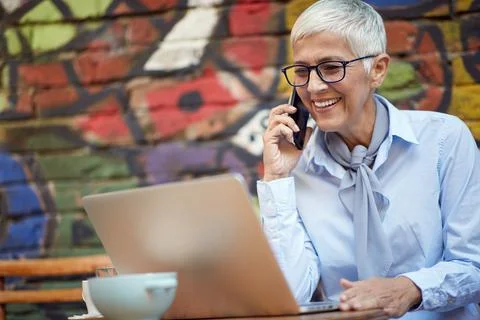 An older woman has a phone call while working on a laptop in a bar. Leisure,  Stock Photos
