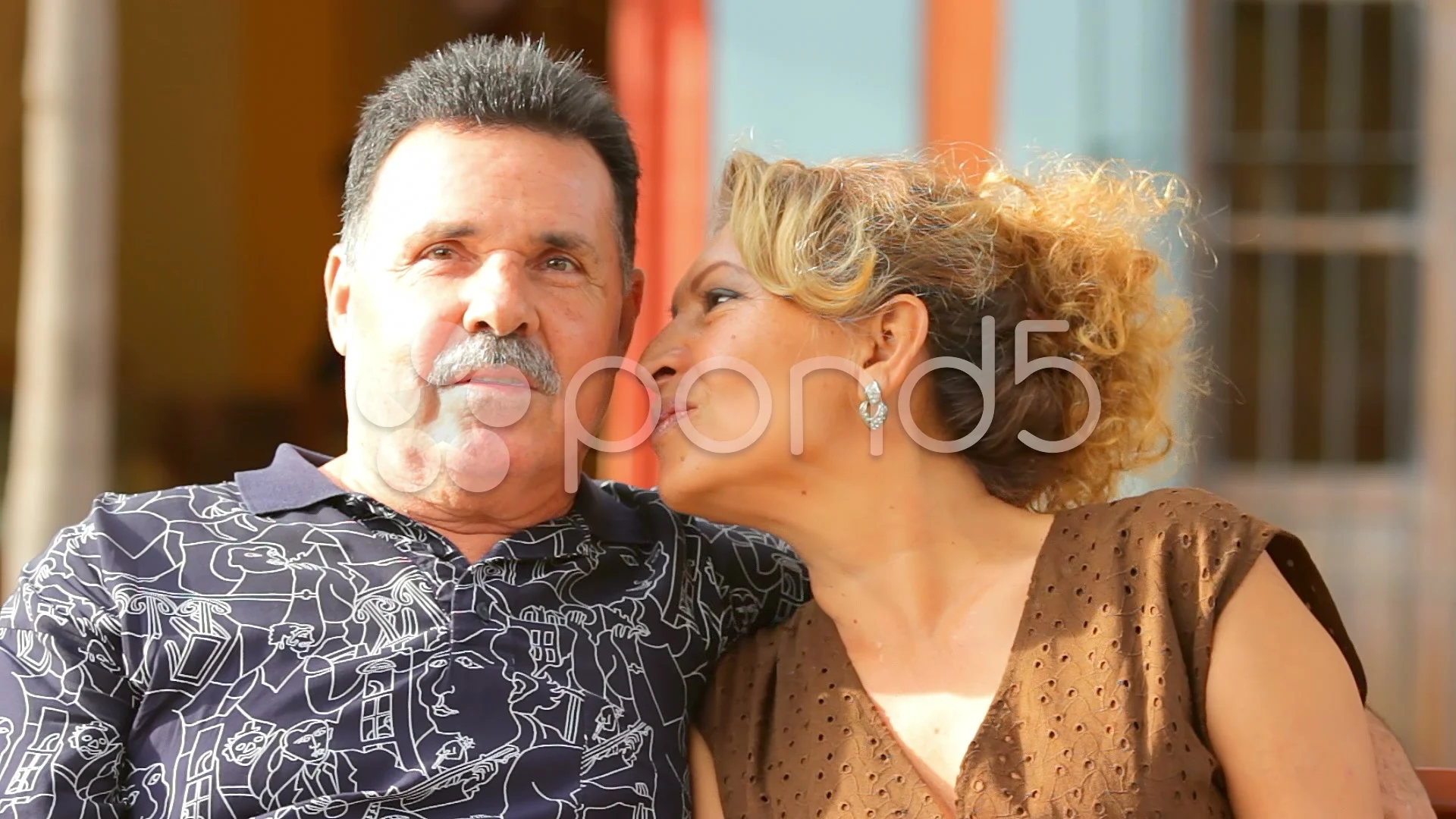 An older woman kisses an older man on the cheek as they sit on a bench