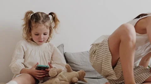 One child plays with a phone while the other is busy with a toy on a sofa in a Vídeos de archivo 336459744