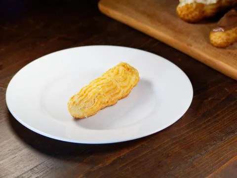One eclair on a white plate, on a dark wooden background. Homemade baking. Stock Photos