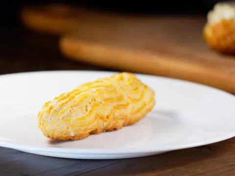 One eclair on a white plate, on a dark wooden background. Homemade baking. Stock Photos