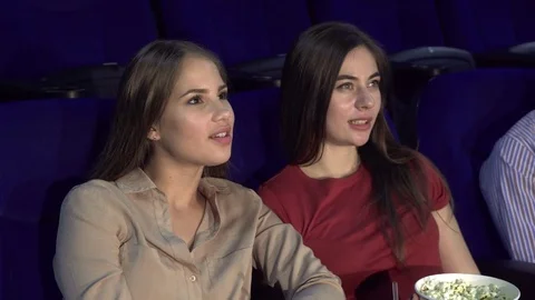 One girlfriend whispers something to a second girl while watching a movie Stock Footage 83941642