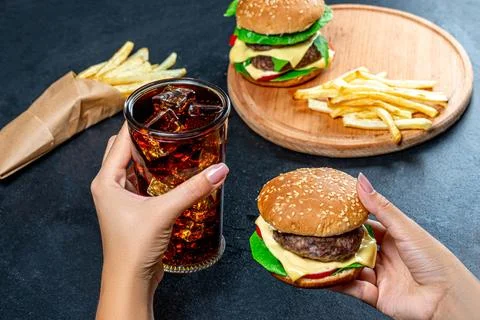 In one hand Burger, in the second the glass with cold Cola. The concept of .. Stock Photos