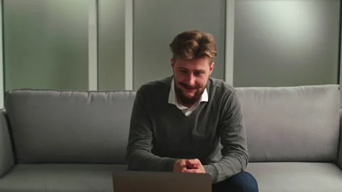 One Handsome Man with Beard is Sitting on the Couch, Using Laptop, Laughing Stock Footage 134796215