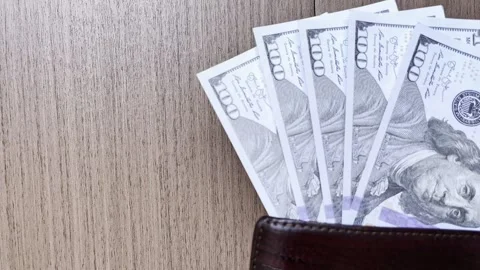 One hundred dollar bills fan out from a brown leather wallet on a wooden table. 스톡 동영상 345557891