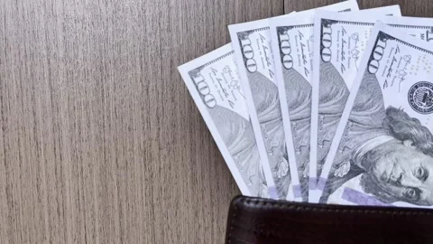 One hundred dollar bills fan out from a brown leather wallet on a wooden table. 스톡 동영상 345557903
