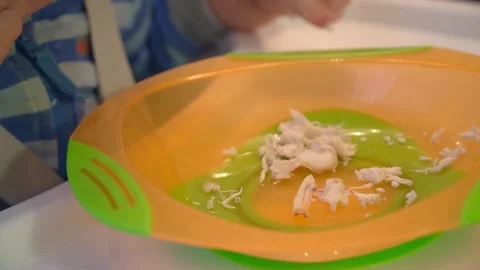 A one-year-old boy eats boiled chicken meat from a plastic plate with his hands. Stock Footage 308029704
