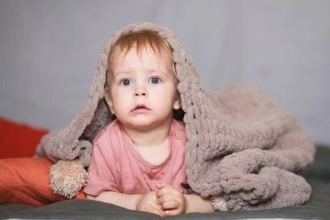 A one-year-old boy hides under a beige knitted blanket or bedspread. Stock Photos
