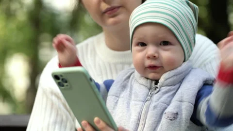 A one-year-old boy sits in his mother's arms in the park and watches a cartoon o Vídeo Stock 210849201