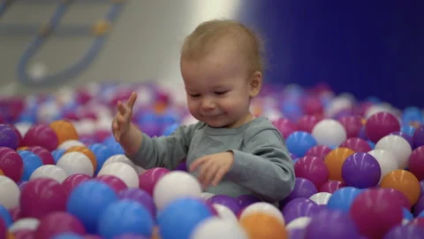 A one-year-old boy sits in a pool with plastic colorful balls and plays with Stock Footage 229985988