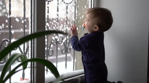A one-year-old boy stands at the window of the house. The child looks out the Stock Footage 231606571