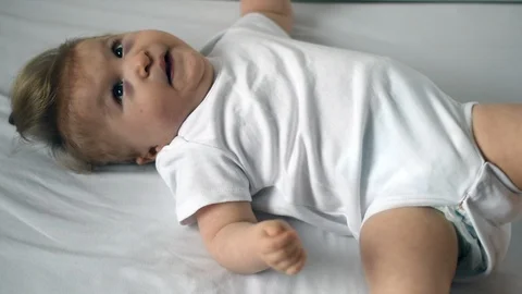 One year old disabled child in white bodysuit is trying to roll on the bed Stock Footage 101794372