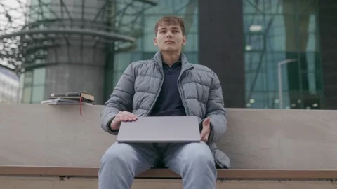 One young man closing laptop and resting on bench near modern glass building. Stock-Footage 332765993