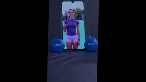 Online personal trainer on a smartphone screen giving a thumbs up with two dumbb Stock Footage 327447730