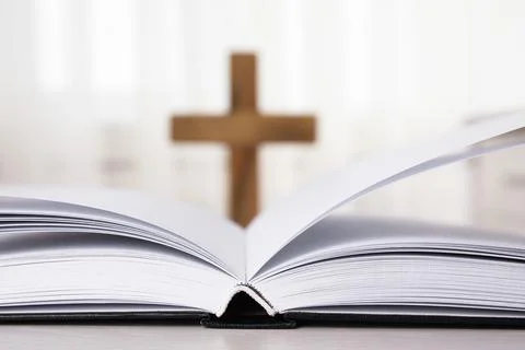 Open Bible on table and blurred cross on background, closeup. Christian relig Stock Photos