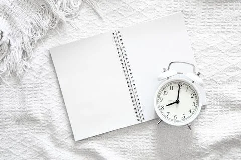 Open blank notepad and white alarm clock in bed, top view. Stock Photos