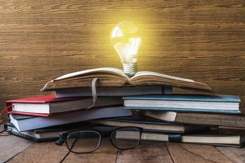 Open book with light bulb, hardback books and glasses on wooden table. Stock Photos