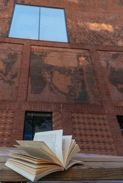 An open book resting on a bench in front of a red brick building Stock Photos