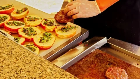 Open buns with herbs and chopped vegetables, and a small burger cooking on the Stock Footage 323915364