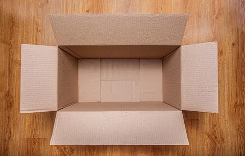 An open, empty cardboard box sits centered on a light brown wooden floor Stock Photos