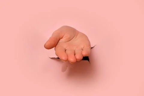 Open hand reaching through torn hole in pastel pink paper background. Minimal Stock Photos