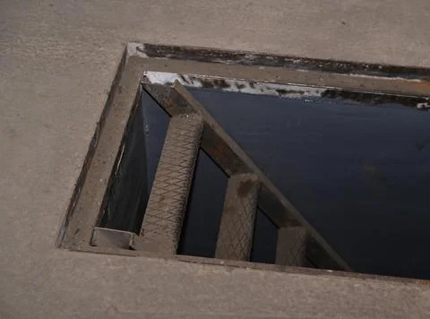 Open hatch leads down via industrial metal stairs into dark basement. Stock Photos