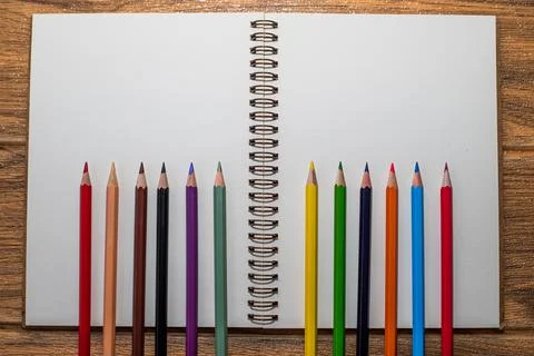 Open notebook with white sheets on the table and many colored pencils lined u Stock Photos