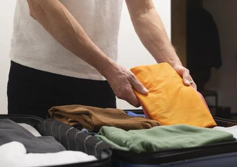 Open suitcase. Cropped hands stack clothing and place essential items into fu Stock Photos