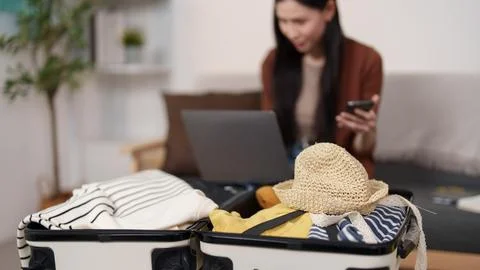 Open suitcase packed with clothes and straw hat with woman using laptop in Stock Photos