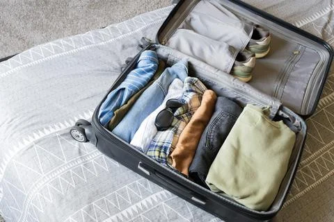 Open suitcase packed with folded clothes, shoes and sunglasses lying on a bed Stock Photos