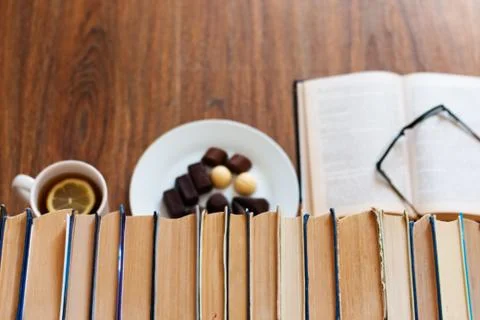 Open textbook, glasses, cup of tea and chocolate candies on white plate, stac Stock Photos