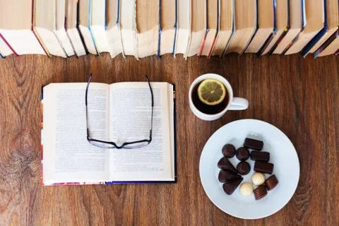 Open textbook, glasses, cup of tea and chocolate candies on white plate, stac Stock Photos