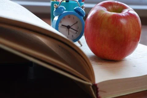 Opened book on the table near the window with an apple and an alarm clock. Stock Photos