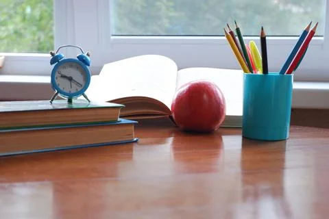 Opened book on the table near the window with an apple and an alarm clock. Stock Photos