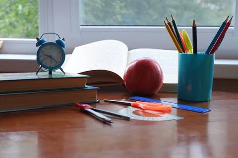 Opened book on the table near the window with an apple and an alarm clock. Stock Photos