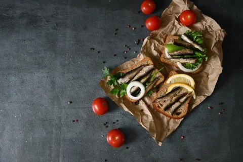 Opened cans with sprats on grey textured stone table with bread, tomatoes and Stock Photos