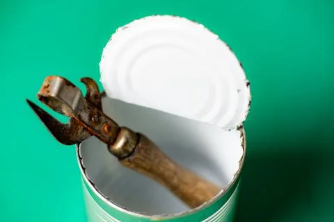 Opened empty tin can and a rusty bottle opener on a green background. Stock Photos
