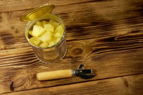 Opened tin can of pineapple pieces and can opener on wooden table Stock Photos