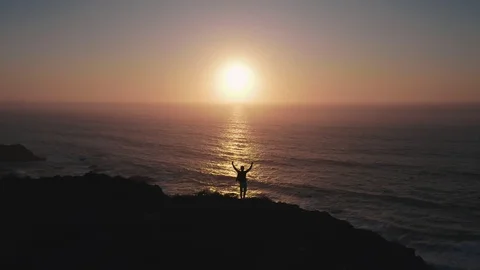 An optimistic bird's eye view of a happy boy on cliff over ocean at sunset Stock Footage 128630457