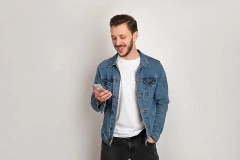 Optimistic hipster in denim casual jacket is looking at his phone smiling. Stock Photos