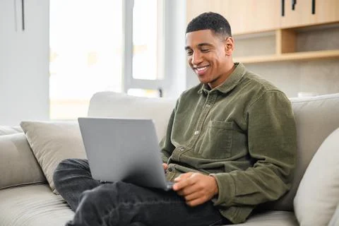Optimistic man in casual wear using laptop while sitting on the couch. Young Stock Photos