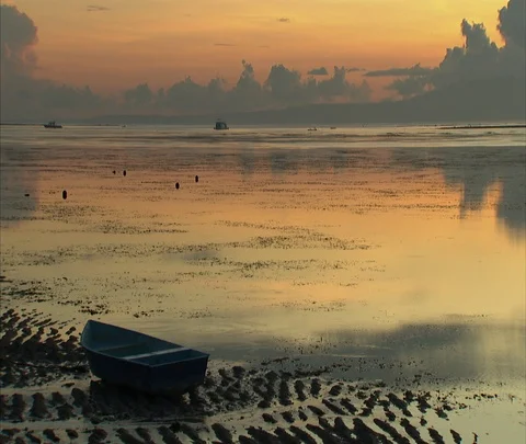 Orange and golden sun reflected in still waters, boat beached in wet sand Stock Footage 86747663