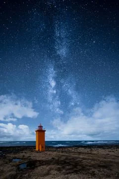 An orange lighthouse by the ocean at night with epic milky way on the sky Stock Photos