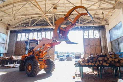 Orange log loader with grapple inside a sawmill warehouse near stacked timber Stock Photos