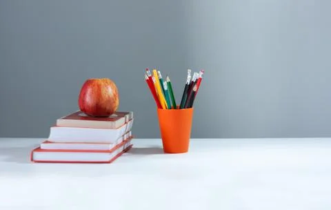 Orange pencil holder, stack of books on white table with red apple. Books sta Stock Photos