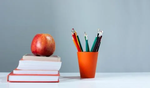 Orange pencil holder, stack of books on white table with red apple. Books sta Stock Photos